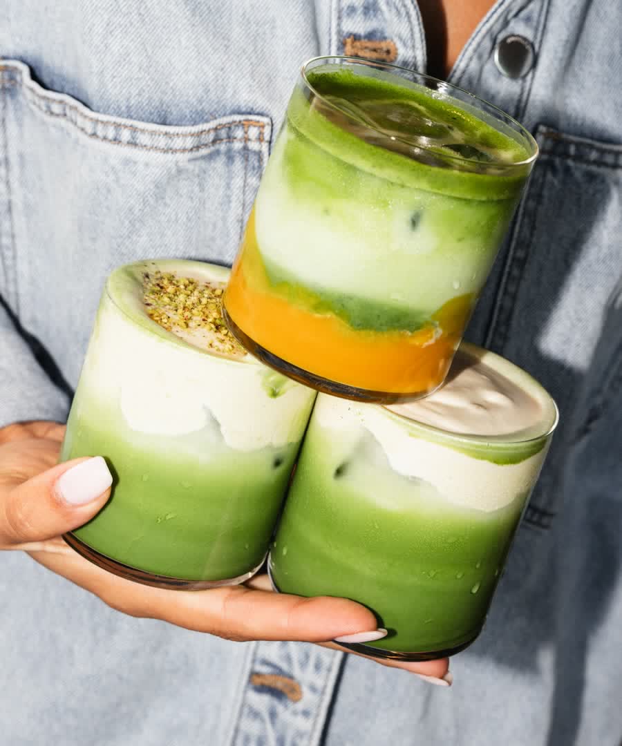 Three layered matcha and pistachio drinks held up in the sun.