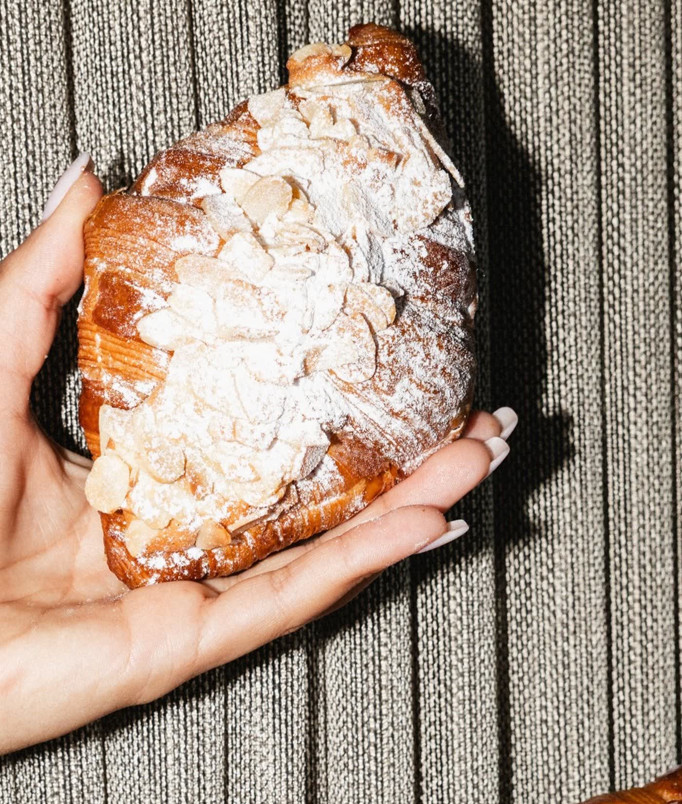 A hand holding a flaked almond croissant above a timber bench at SANI Bakehouse.